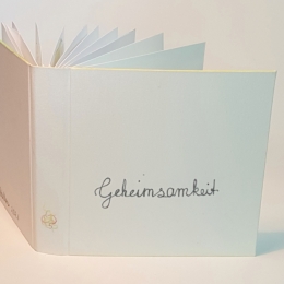 Blick auf den weißen Einband eines offenen Buches. Unten auf dem Buchrücken ist buntes Krikelkrakel. Die Kanten des Einbands sind gelb, rot und grün angemalt. Auf dem Cover steht in Schreibschrift „Geheimsamkeit“.  Die nächsten Bilder zeigen je eine Doppelseite des Buches mit einer Gedichtzeile.