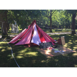 Bild zeigt eine Art Zelt aus einzelnen Stoffbahnen in Rottönen.  Das Zelt steht im Grünen. Vor dem Zelt liegt ein Teppich. Daneben lehnt ein hölzernes Schild.