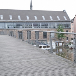 Eine Brücke erstreckt sich in einem flachen Bogen über die Fulda. In der Mitte der Brücke sind Sitzbänke.  Der Brückenboden ist aus Holz. Im Brückengeländer aus Maschendraht sind lange, schmale, dunkelblaue Streifen zu erkennen. Sie sind alle gleich lang und in unregelmäßigem Abstand im Geländer befestigt. sie fallen kaum auf.