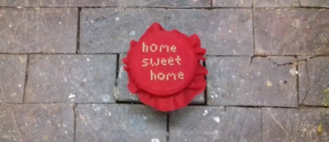 Ein Schraubglas mit rotem Stoffdecken darauf. Es ist im Keuzstich mit dem Schriftzug "home sweet home" bestickt. Es steht auf einem Boden aus Holzklötzern.