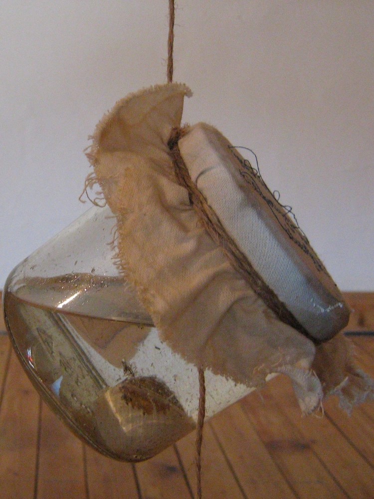 Link zur Installation "this would be a safe space". Ein Weckglas, dessen Deckel mit einem Stück Stoff bezogen ist. Das Glas hängt an einem Faden und im Glas sind eine klare Flüssigkeit und ein Schneckenhaus mit großem Loch, eingeätzten Loch.