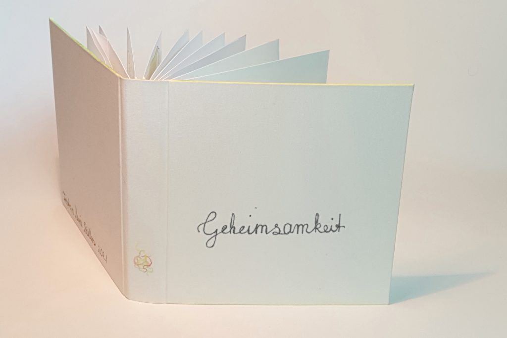 Link zum Buch "Geheimsamkeit". Blick auf den weißen Einband eines offenen Buches. Unten auf dem Buchrücken ist buntes Krikelkrakel. Die Kanten des Einbands sind gelb, rot und grün angemalt. Auf dem Cover steht in Schreibschrift "Geheimsamkeit".