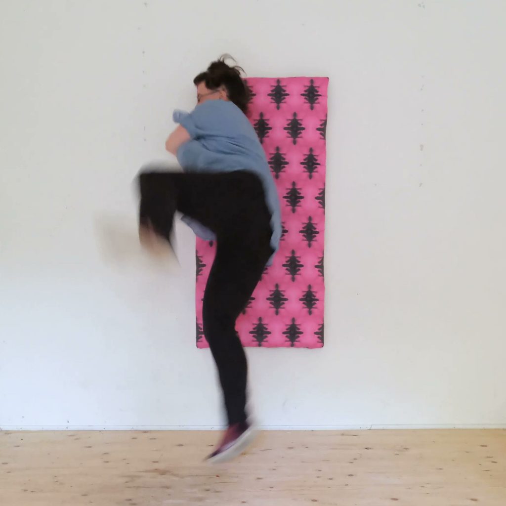 Link zu Videos von den "WTFX.performances". Es gibt keine Audiodeskription. Das Still aus einer Performance zeigt einen Raum mit Holzboden und hellen Wänden. Mittig an der Wand hängt ein gepolstertes Brett. Es ist Pink und Auberginefarben gemustert. Tristan Marie springt dagegen. Nin trägt eine schwarze Hose und ein hellblaues Shirt. 