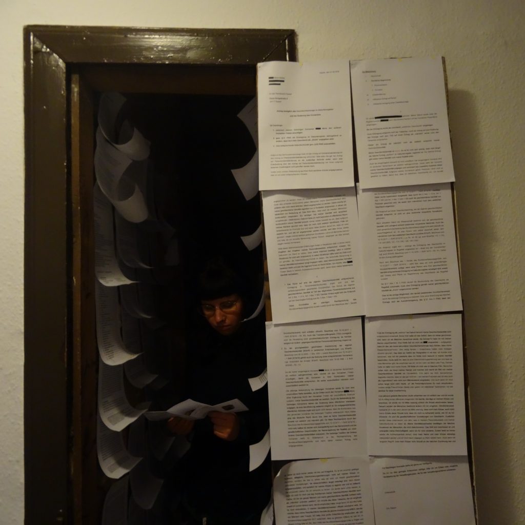 Link zur Installation "Das ist nicht mein Platz". Die offene Tür zu einer winzigen Abstellkammer. Tür und Wände sind mit bedrucktem Papier behangen. Eine Person kommt aus der dunklen Kammer heraus.