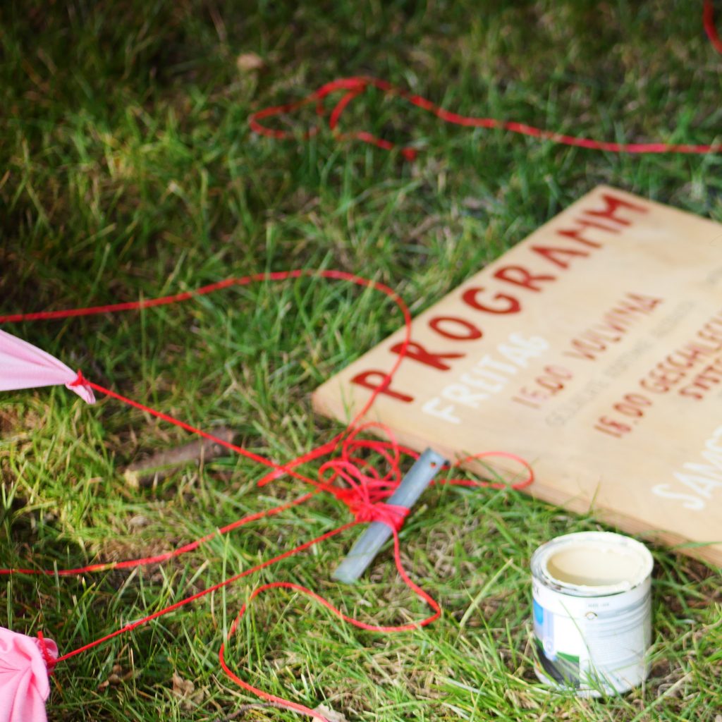 Link zur Arbeit "Rotes Zelt". Auf einer grünen Wiese liegt ein hölzernes Schild mit der Aufschrift "Programm". Daneben sind einige Zeltspannschnüre und kleine Ecken des Zeltes in Rosatönen zu sehen.