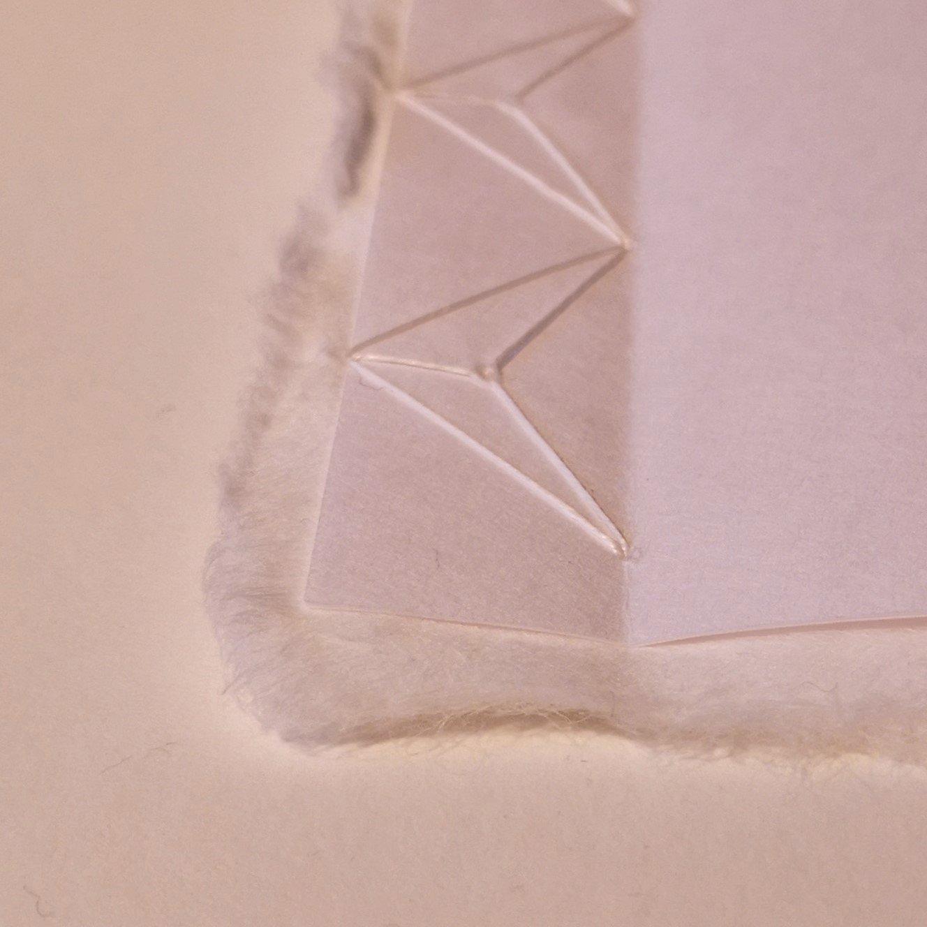 Detailbild: Auf weißem Grund liegt ein schmales Heft aus weißem Papier. Es ist mit weißem Zwirn in japanischer Bindung gebunden. Der Einband ist aus recht zartem Papier. Die Buchseiten sind aus hauchdünnem Papier mit Büttenrand. Er schaut wie Fell am Buchrücken und der oberen und unteren Kante des Buches hervor.